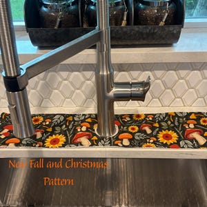 May include: A rectangular fabric sink splash guard featuring a black background with a repeating pattern of orange mushrooms, yellow sunflowers, and green leaves. The mat is positioned around the base of a stainless steel kitchen faucet.