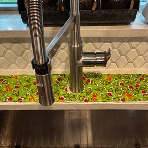 May include: A rectangular fabric sink splash guard featuring a repeating pattern of green cartoon characters wearing red Santa hats on a bright green background with white star accents and the text Feelin Grinchy printed in red and yellow letters.
