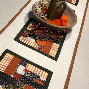 Peut inclure: Un chemin de table matelassé avec un motif patchwork de tissus orange, blanc et noir, incluant des imprimés de citrouilles et de carreaux. Un bol en bois contient un vase en verre vert olive mat, des rubans orange et des tiges de baies blanches.