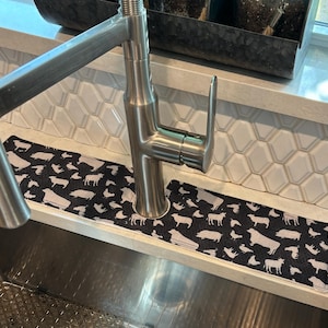 May include: A rectangular black fabric sink splash guard featuring a repeating white pattern of various farm animals including cows, pigs, and chickens. The mat has a central circular cutout designed to fit around a stainless steel kitchen faucet.