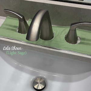 May include: A light sage green fabric faucet absorbent mat featuring three circular cutouts for a bathroom sink faucet and two handles. The mat is positioned around a brushed nickel faucet assembly. The text Color Shown: (Light Sage) is visible.