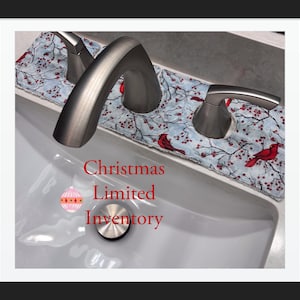 May include: A fabric faucet absorbent mat with a light blue background, red cardinal birds, and berry-covered branches, positioned around a silver bathroom faucet. The text Christmas Limited Inventory is displayed in red with a pink ornament graphic.