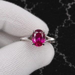 Puede incluir: Un anillo de plata con una vibrante piedra preciosa fucsia de corte ovalado sujeta por cuatro garras. La banda tiene un diseño de vástago dividido que se estrecha hacia la piedra central, mostrando un acabado metálico pulido y una gema facetada.