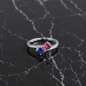 May include: A silver-toned bypass ring featuring two round-cut stones, one deep blue and one vibrant pink, set in a polished metal band. The design showcases a crossover style where the two stones are positioned side-by-side at the centre of the ring.