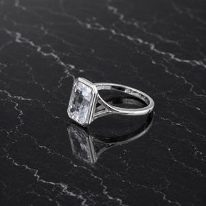 May include: A silver-toned ring featuring a large, rectangular, emerald-cut clear stone set in a bezel setting. The band has a split-shank design that connects to the stone setting, creating a polished and reflective metallic appearance throughout.