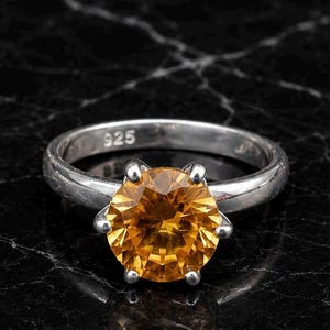 Peut inclure: Une bague en argent avec une pierre précieuse ronde, facettée et jaune doré, maintenue par un sertissage à six griffes. L'intérieur de l'anneau porte clairement l'inscription 925, indiquant une construction en argent sterling au fini poli.