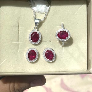 Puede incluir: Un conjunto de joyería que consta de un collar plateado con un colgante ovalado rojo, un anillo a juego con una piedra ovalada roja y dos pendientes de botón ovalados rojos. Cada pieza presenta un halo de pequeñas piedras blancas.