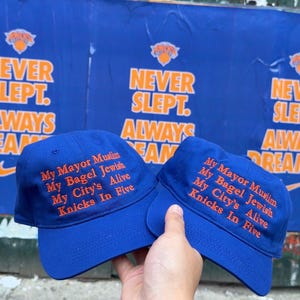 Op de afbeelding: Twee koningsblauwe baseballpetten met oranje geborduurde tekst: My Mayor Muslim, My Bagel Jewish, My City's Alive, Knicks In Five. De petten hebben een gestructureerde kroon en een gebogen klep in een opvallende kleurencombinatie.