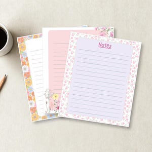 May include: Three rectangular stationery sheets with lined writing areas. The top sheet is light purple with the word Notes printed at the top and a pink floral border. The middle sheet is pink with floral accents. The bottom sheet has a colorful floral border.