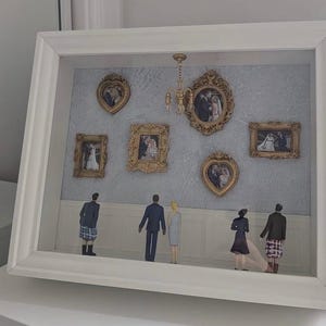 May include: A white shadow box frame containing a miniature gallery scene with five ornate gold frames displaying wedding photographs, a small gold chandelier, and five miniature figures standing on a white base looking at the wall.
