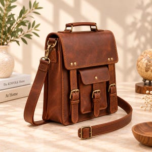 May include: A brown leather satchel with a top handle and an adjustable shoulder strap. The bag features a fold-over flap with two buckle closures, a front pocket with a buckle, and antique brass-toned metal hardware throughout the design.