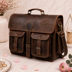 May include: A dark brown leather satchel featuring a top handle, a shoulder strap, and two front pockets with buckle closures. The front flap is embossed with the name JOHN and a compass rose design. The bag includes antique brass hardware.