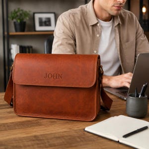 May include: A rectangular, cognac-coloured leather messenger bag with a front flap featuring the embossed name JOHN. The bag includes a matching adjustable shoulder strap attached to the sides with metal hardware and has a smooth, structured finish.