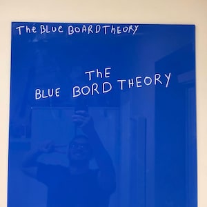 May include: A rectangular blue glass dry-erase board with a reflective surface. The text "The BLUE BOARD THEORY" is written in white marker at the top, and "THE BLUE BORD THEORY" is written in larger white letters in the centre of the board.