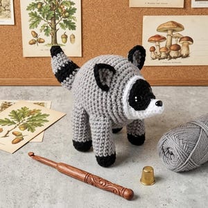 May include: A handmade crochet raccoon plush toy featuring a round grey body, a striped tail, black ears, and a black and white face mask with dark eyes. The raccoon is positioned near a wooden crochet hook, a gold thimble, and a ball of grey yarn.