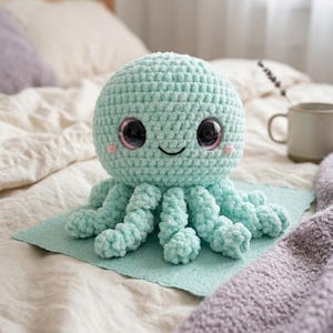 May include: A mint green crocheted octopus plush toy with a round head, eight curled tentacles, large dark glossy eyes, and small pink embroidered cheeks. The soft yarn texture is visible throughout the stuffed creature's rounded body.