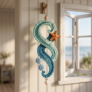 May include: A decorative wall hanging featuring a crocheted S-shaped wave in shades of light blue, teal, and dark blue. The piece is accented with an orange starfish, two small light blue crocheted spheres, and a single white bead at the bottom.