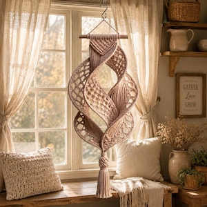 Puede incluir: Un tapiz de macramé beige con un complejo diseño de espiral retorcida y patrones de nudos intrincados. La pieza cuelga de un soporte de madera horizontal mediante un gancho metálico y termina en una borla gruesa y anudada en la parte inferior.