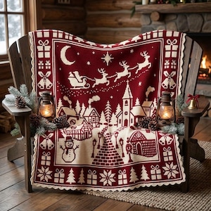 Op de afbeelding: Een rechthoekige gebreide deken met een kerstdorp-tafereel in donkerrood en crème. Het ontwerp bevat een arrenslee met rendieren, een maansikkel, een kerk, huizen, een sneeuwpop, cadeauverpakkingen en klokken.