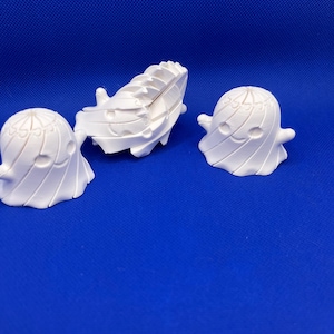 May include: Three small, white, three-dimensional ghost figurines with smooth, rounded surfaces. Each ghost features a simple, carved facial expression with two circular eyes and a curved mouth, along with small, outstretched arms on either side.