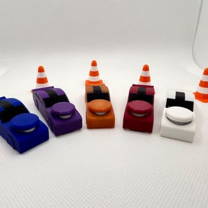 May include: Five small toy race cars in blue, purple, orange, red, and white, each featuring a black roof stripe and a circular button on the hood. The cars are arranged in a row, interspersed with six miniature orange and white traffic cones.