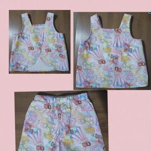 May include: A sleeveless top with a split back design and matching shorts, both featuring an all-over print of Barbie dolls in various gowns and the Barbie logo in pink script against a light, multi-coloured background.