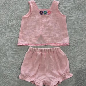 May include: A light pink sleeveless top and matching shorts set made of textured fabric. The top features three small embroidered flowers in purple, blue, and pink at the neckline and a decorative zigzag stitch along the bottom hem.