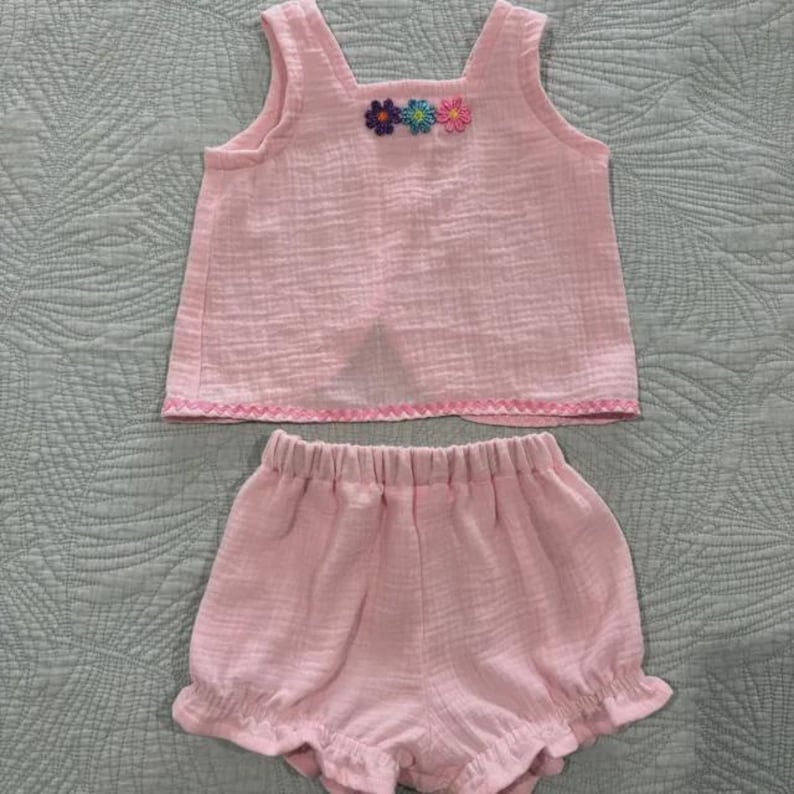 May include: A light pink sleeveless top and matching shorts set made of textured fabric. The top features three small embroidered flowers in purple, blue, and pink at the neckline and a decorative zigzag stitch along the bottom hem.