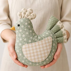 Peut inclure: Un jouet poule en tissu rembourré avec un corps vert sauge à motif floral blanc, une aile à carreaux vichy beige et blanc, un bec jaune moutarde, ainsi qu'une crête et une queue en tissu vert à pois blancs et détails floraux.
