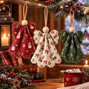 Può includere: Tre angeli decorativi in tessuto appesi a un ramo. L'angelo a sinistra è rosso con pupazzi di neve, quello centrale è color crema con fiocchi e pigne, e quello a destra è verde con fiocchi di neve dorati. Ognuno ha una testa in legno.
