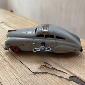 Puede incluir: Un coche de juguete gris vintage con ruedas rojas y una llave de cuerda. El coche tiene un diseño aerodinámico con el texto "Auto Tex" en la parte superior y "Schuco" en la llave. El juguete está sobre una superficie de madera.
