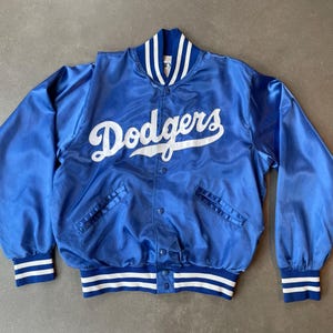 Könnte beinhalten: Eine blaue Satin-Collegejacke mit dem Schriftzug Dodgers in weißer Schreibschrift auf der Brust. Die Jacke verfügt über eine Druckknopfleiste, lange Ärmel sowie gerippte Bündchen an Ärmeln und Saum mit weißen Streifen. Der Kragen ist ebenfalls gerippt.