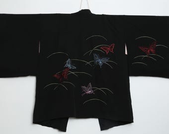 May include: Black kimono with embroidered butterflies in red, blue, and purple. The butterflies are set against a backdrop of curved white lines. The kimono is hanging against a white background.