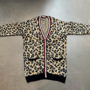 Può includere: Un cardigan con stampa leopardata con scollo a V, finiture nere e due tasche frontali. Il maglione ha un bordo a righe rosse, bianche e nere lungo lo scollo. Le maniche hanno polsini neri.