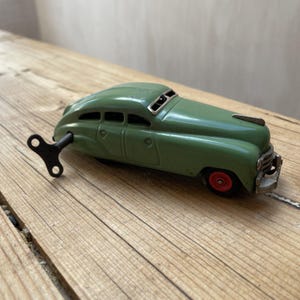May include: A vintage green wind-up toy car with red wheels and a silver hood ornament. The car has a key attached. The toy car is set against a wooden background. A classic collectible.