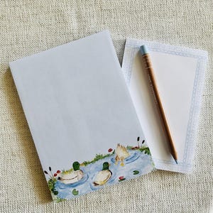 May include: A rectangular notepad featuring a watercolor illustration of three ducks swimming in a blue pond at the bottom, accompanied by a smaller notepad with a light blue patterned border and a brown wooden pencil resting on top.