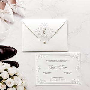 May include: A white wedding invitation set featuring an envelope with a pearl closure and a monogram of the letters M and K. The invitation card displays the names Mira and Kenan, event details, and ornate embossed borders on textured white paper.