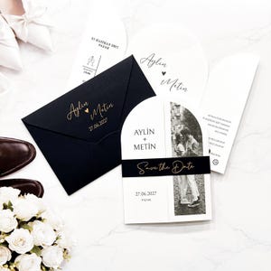 May include: A black envelope with gold text reading Aylin Metin 27.06.2027, alongside arch-shaped white wedding invitation cards. One card features a black band with the text Save the Date, a black and white photograph of a couple, and the names Aylin + Metin.