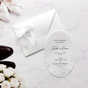 Könnte beinhalten: Eine ovale, weiße Hochzeitseinladungskarte mit geprägtem Blumenrand und schwarzem Text, der Selda & Kaan lautet, begleitet von einem passenden weißen Umschlag mit weißer Schleife und den geprägten Initialen S/K, neben weißen Schuhen und Rosen.