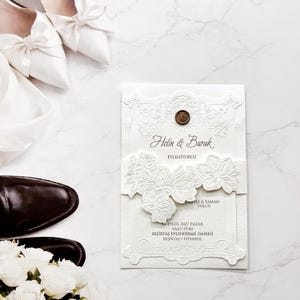 May include: A white wedding invitation card featuring embossed floral details, a circular wax seal, and text including Helin & Burak, EVLENIYORUZ, 12 EYLÜL 2027 PAZAR, SAAT:19:00, BEŞIKTAŞ EVLENDIRME DAIRESI, BEŞIKTAŞ / ISTANBUL, alongside white shoes and flowers.