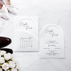 Peut inclure: Un ensemble de faire-part de mariage blanc en forme d'arche avec les prénoms Ceyda et Tarik en écriture noire. La carte de gauche affiche un calendrier de juin 2027 avec un cœur autour du 27. La carte de droite détaille l'événement au Swissotel.