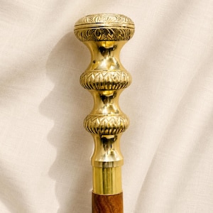 Può includere: Un bastone da passeggio con impugnatura in metallo color oro lucido e dettagli incisi. L'impugnatura presenta una forma arrotondata a più livelli con due sezioni decorative bulbose. L'asta è realizzata in legno marrone scuro venato.