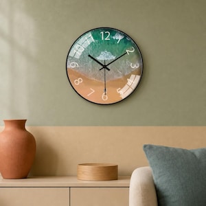 Puede incluir: Un reloj de pared redondo con marco negro y una esfera que muestra una vista aérea de una playa de arena y olas de mar turquesa. El reloj tiene números blancos del uno al doce y finas manecillas negras de hora y minuto.