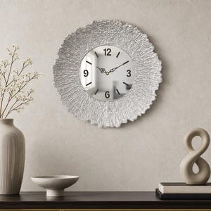 May include: A round wall clock with a silver, textured, radiating frame surrounding a circular mirror face. The clock features black numerals at the 3, 6, 9, and 12 o'clock positions, with black hour and minute hands centered on the reflective surface.
