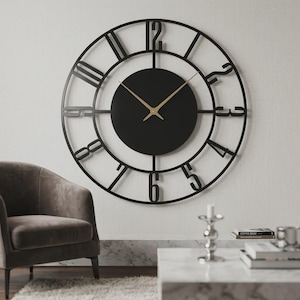 Puede incluir: Un gran reloj de pared circular con un marco de metal negro y números arábigos calados. El centro consta de una esfera circular negra sólida con manecillas finas de color dorado para las horas y los minutos, con un diseño esquelético.