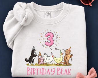 May include: A white crewneck sweatshirt featuring a graphic of a black and white cat, an owl, a chicken, a duck, and a bear wearing party hats. A pink balloon shaped like the number 3 floats above. The text reads BIRTHDAY BEAR.