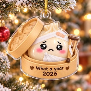 May include: A clear acrylic Christmas ornament featuring a cartoon dumpling inside a bamboo steamer basket. The dumpling has a sad face, pink cheeks, a bandage on its head, and a white arm sling. It holds brown crutches. Text reads: What a year 2026.