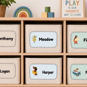 Puede incluir: Seis cajas de almacenamiento rectangulares de tela beige dispuestas en una estantería de madera. Cada caja tiene una etiqueta personalizada con una ilustración de animal única y un nombre: Anthony, Meadow, Finley, Logan, Harper y Noah.