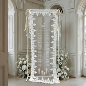 Puede incluir: Un letrero rectangular alto de acrílico transparente para asientos de boda, sostenido por un marco de metal blanco. El letrero presenta texto blanco que dice "Our Favorite People" y enumera ocho mesas, con encaje blanco y cintas beige.