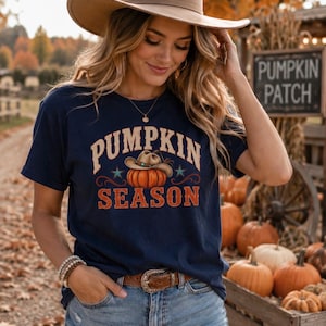 Può includere: Una t-shirt a maniche corte blu navy con una stampa grafica di una zucca arancione che indossa un cappello da cowboy, circondata dalla scritta PUMPKIN SEASON in un carattere curvo. Il design include piccoli accenti a forma di stella.